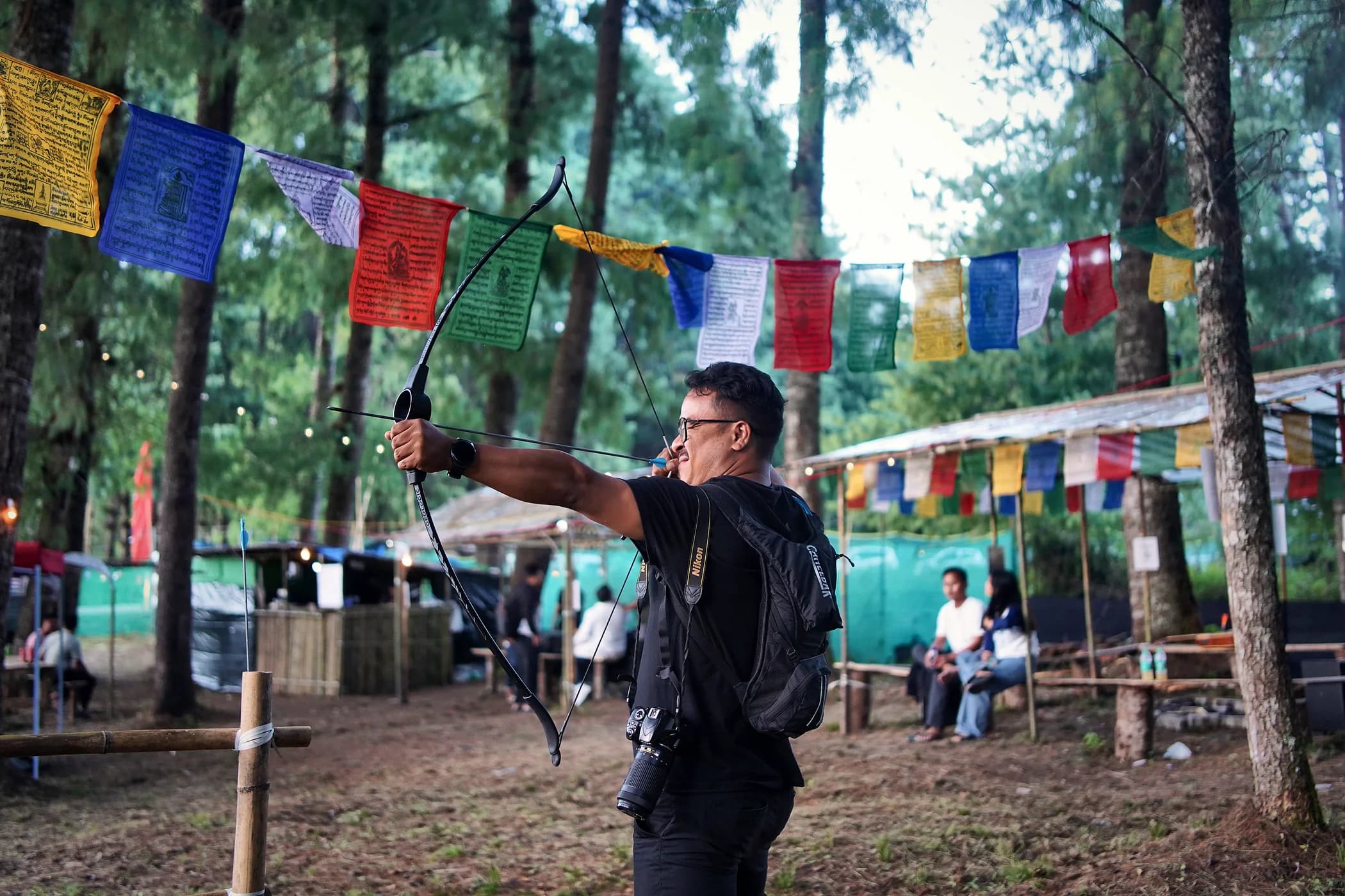 A camper drawing a recurve bow at the camp's archery range, prayer flags strung through the pines