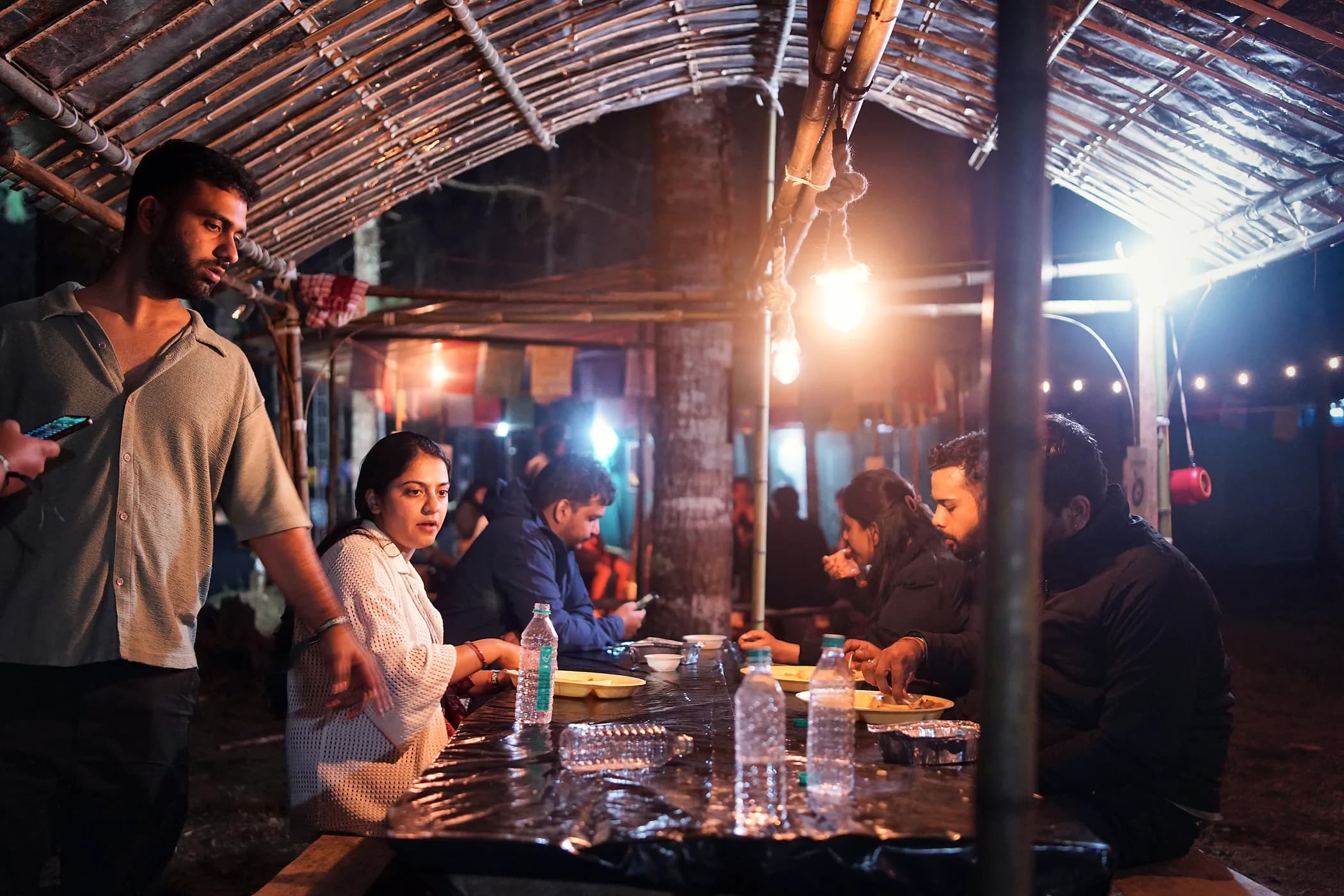 Campers eating at a long table under a bamboo roof, hung with bare bulbs