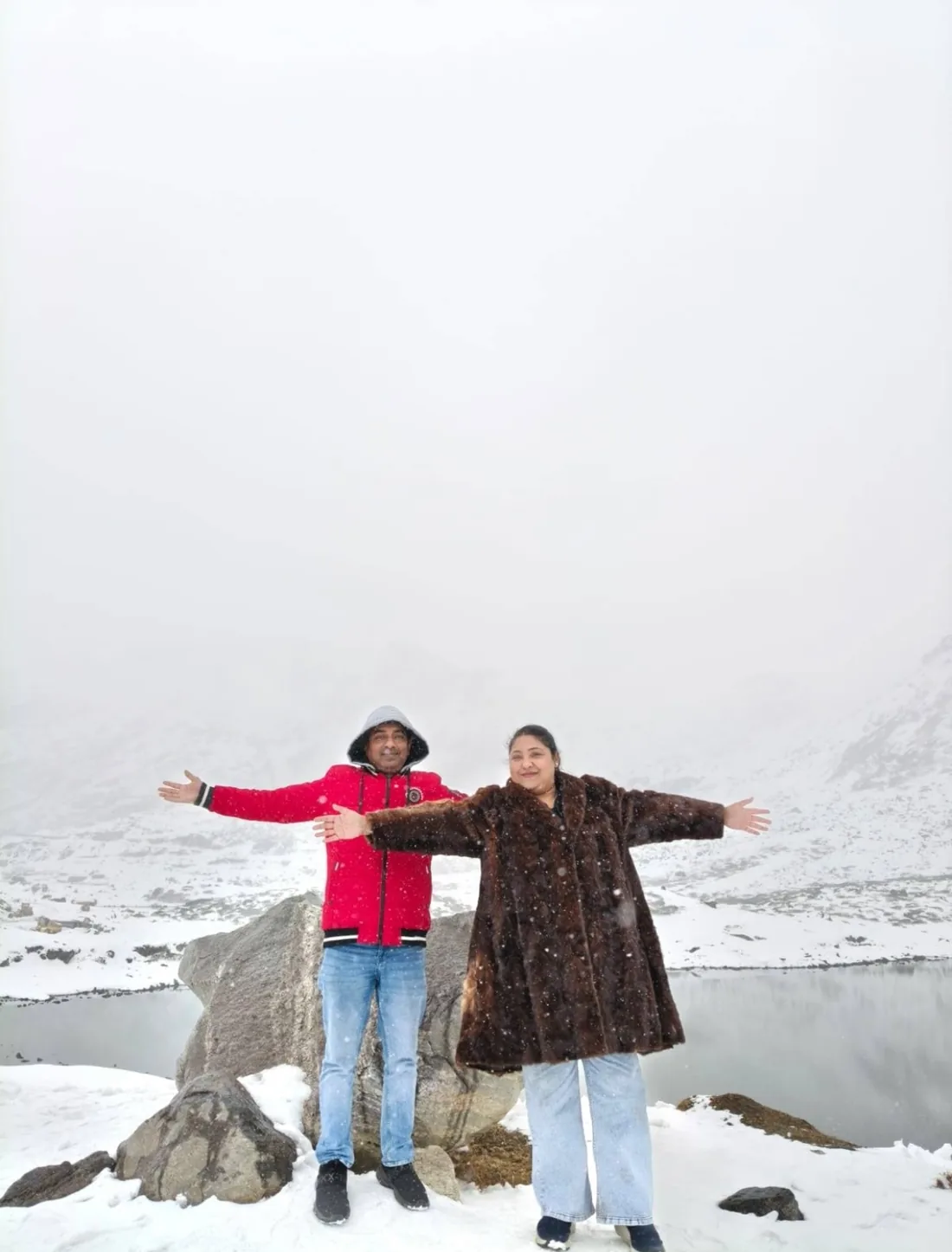 A couple with both arms flung wide beside a boulder, snow falling over the lake and white slopes behind them