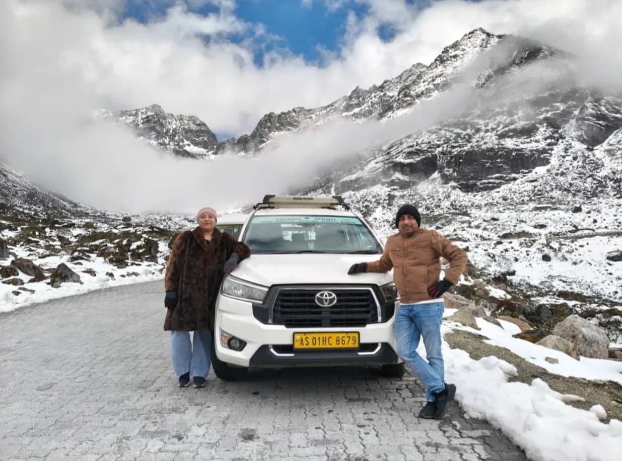 A couple standing either side of their white Innova on a paved mountain road, snow-covered peaks and cloud behind