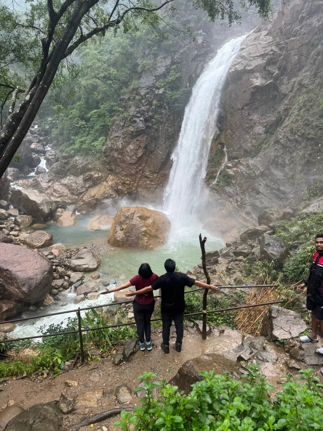A couple at a railing looking across at a tall single-drop waterfall falling past a large pale boulder