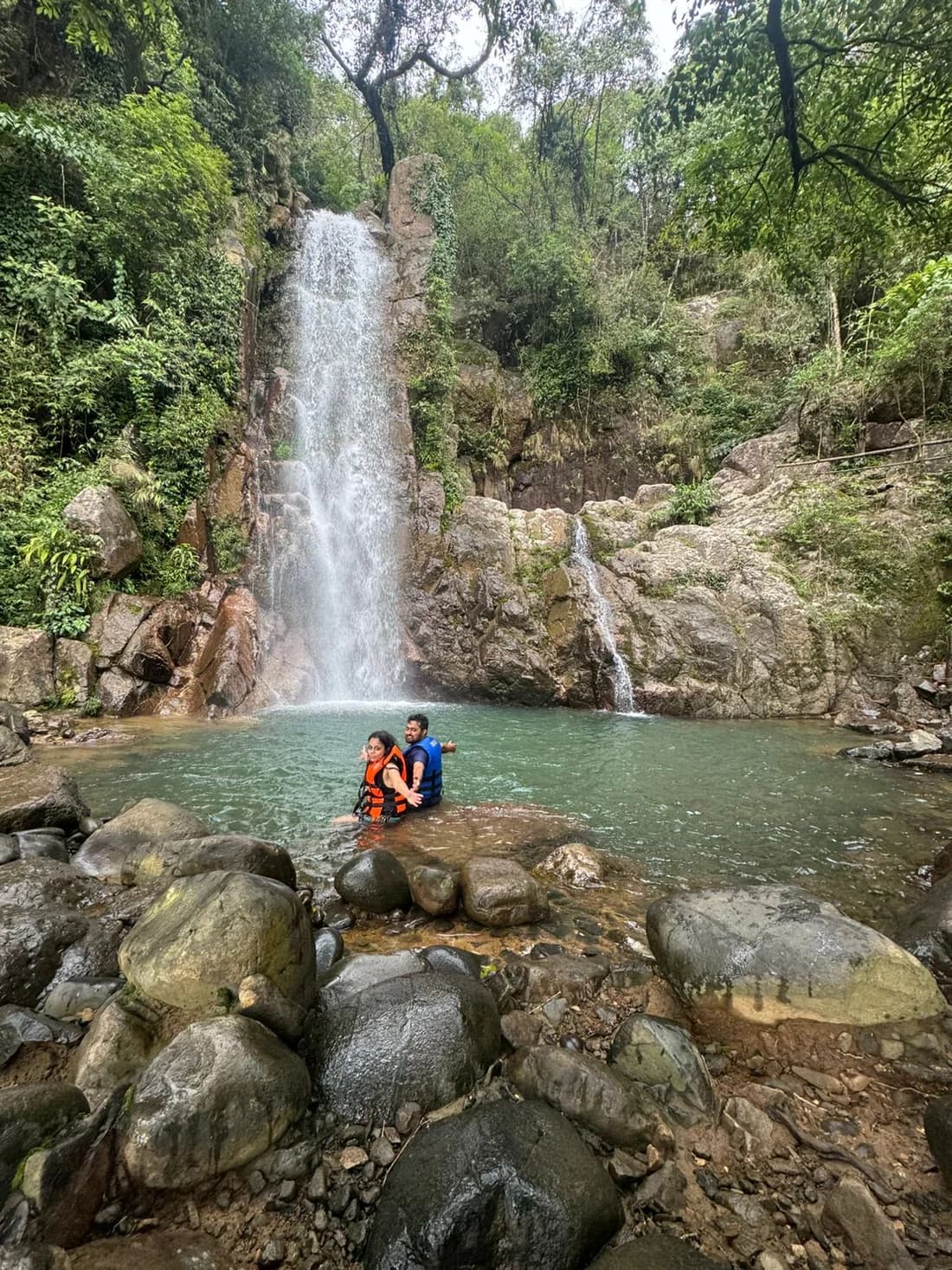 A couple in life jackets sitting in a plunge pool below a tall, narrow waterfall, boulders in the foreground