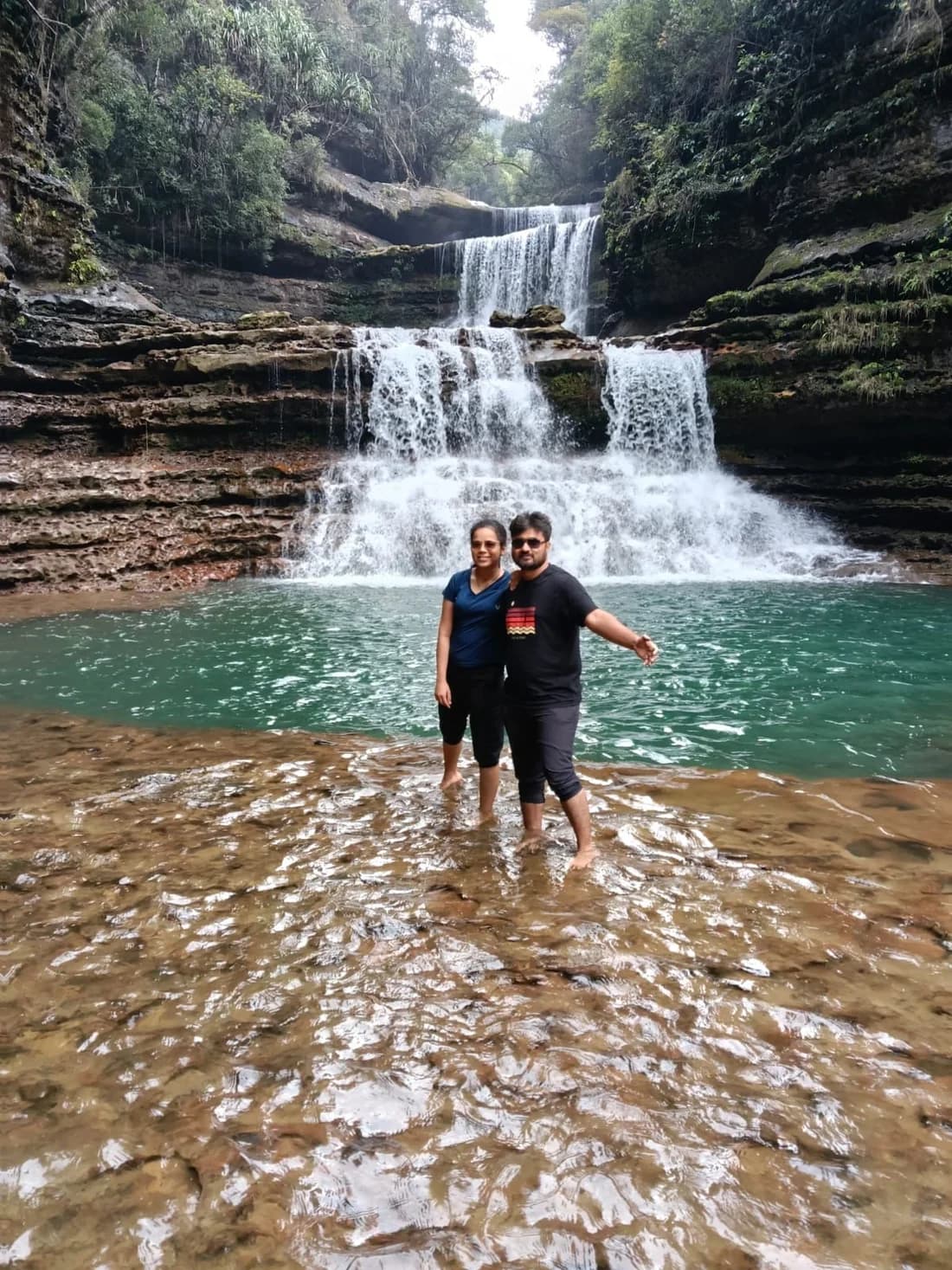 A couple standing together on the rock shelf below a tiered waterfall, the pool turquoise behind them