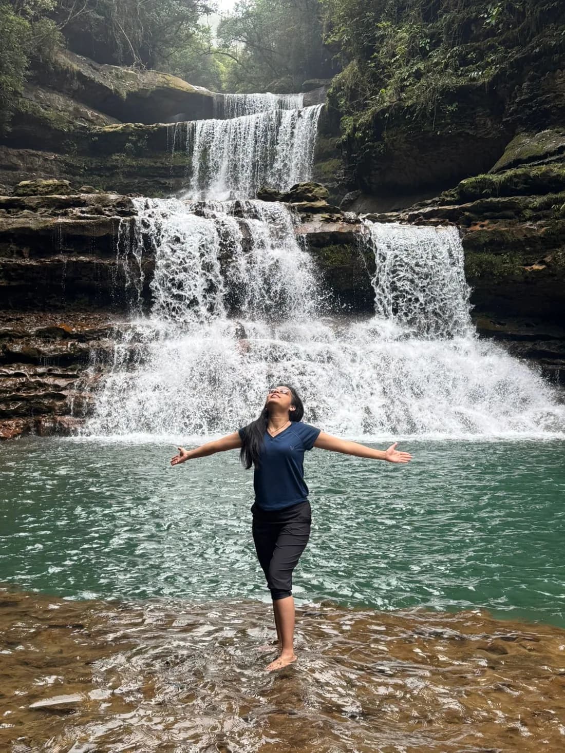 A traveller standing in the shallows with both arms flung wide below a wide, three-tiered waterfall and a green pool
