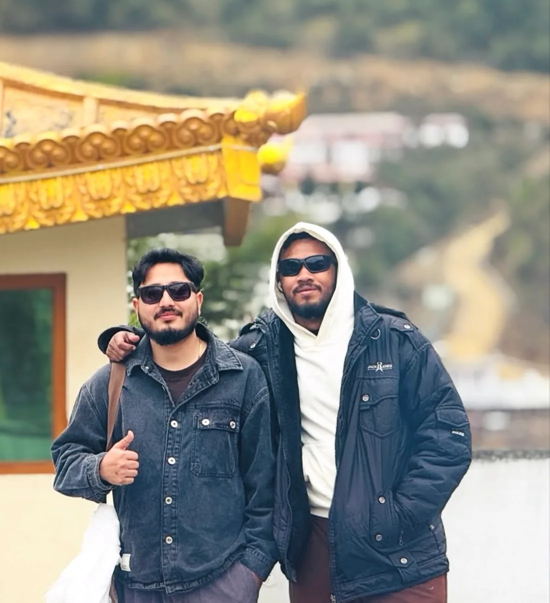 Two travellers in sunglasses in front of a gold-eaved monastery building, a hill road winding away behind them