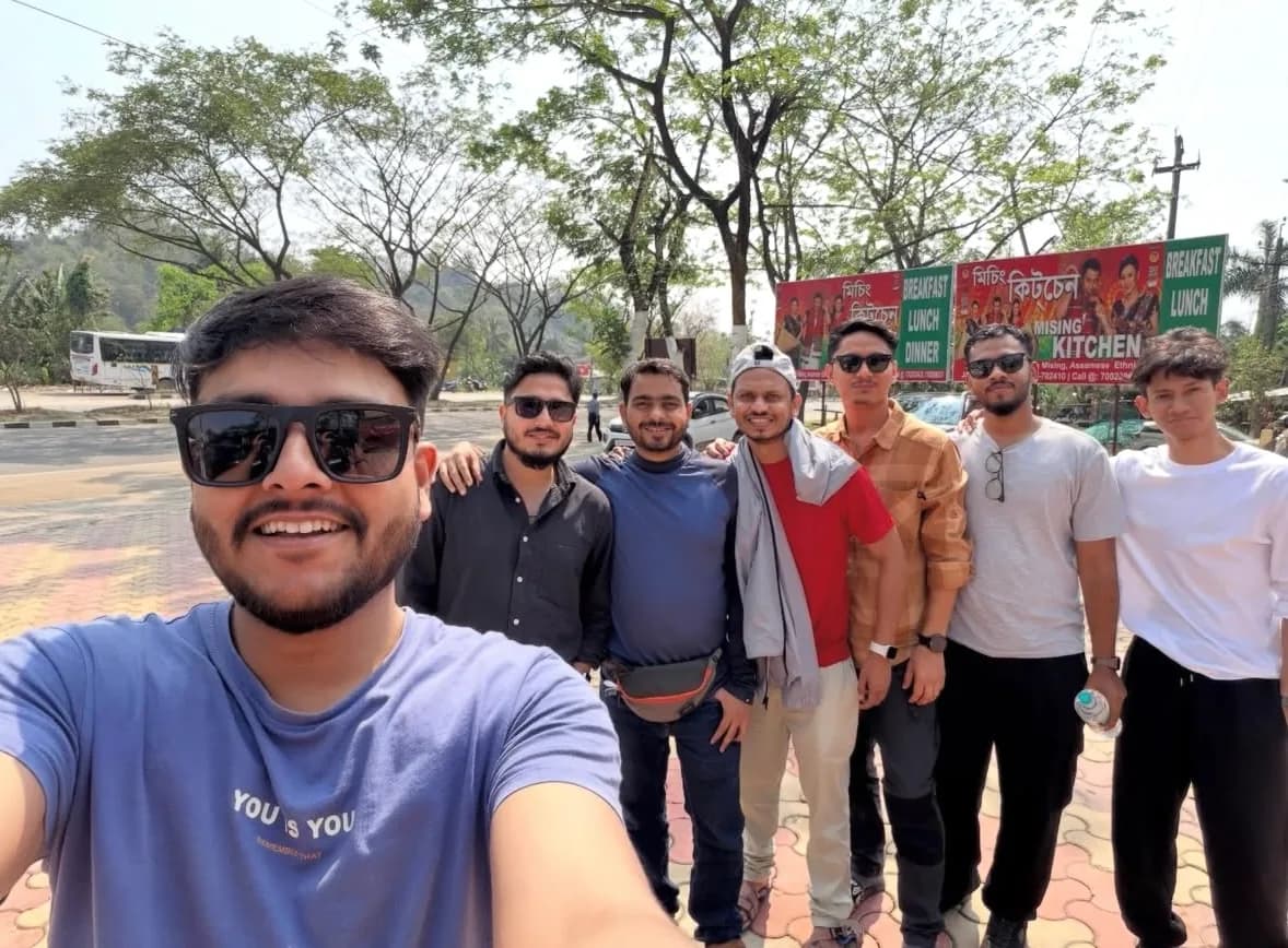 Seven travellers crowded into a selfie at a roadside stop, a Mising kitchen signboard and the highway behind them