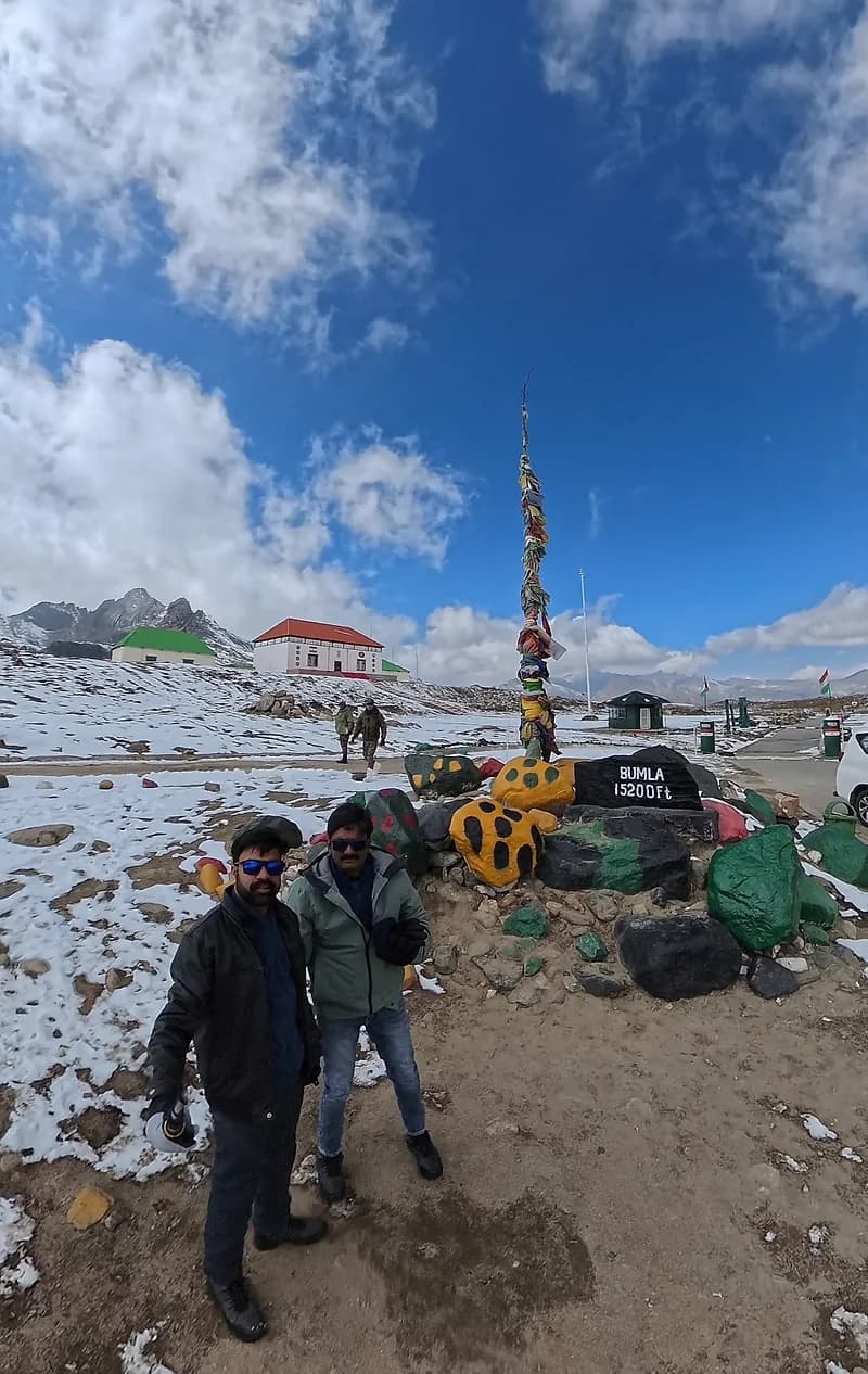 Two travellers beside the Bumla 15,200 ft marker stone, prayer flags on the pole behind