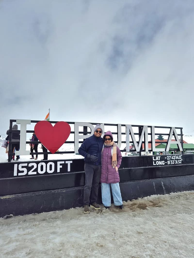 Two travellers at the I ♥ Bumla sign at 15,200 feet, snow underfoot and cloud behind