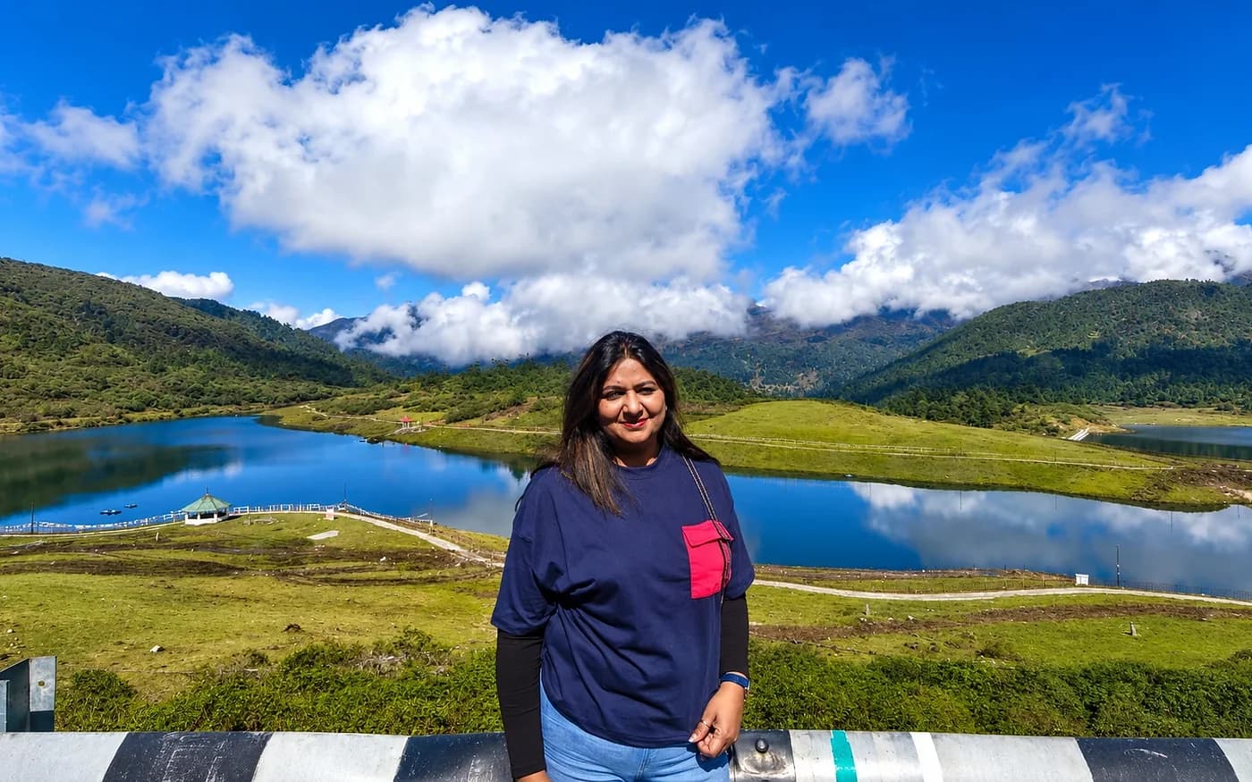 A traveller at a railing above a still lake ringed by green meadow and forested hills