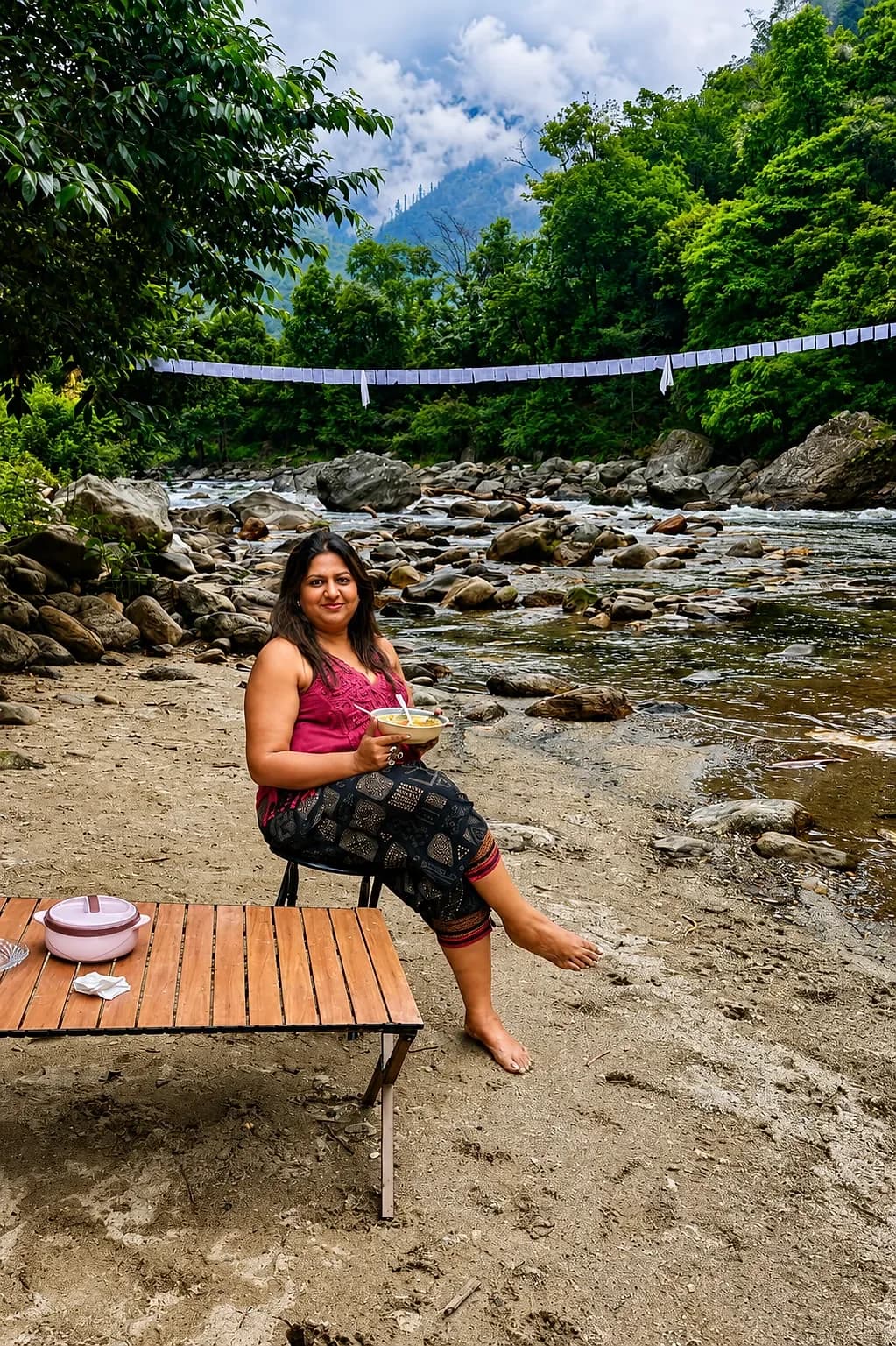 A traveller with a bowl of lunch at a camp table on a river beach, prayer flags strung across the water
