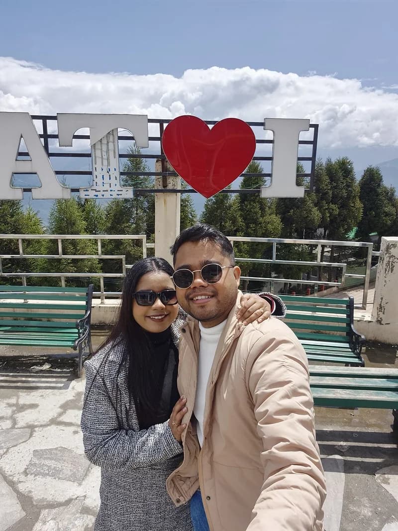 A couple at the Tawang sign, pine forest and cloud over the valley behind them
