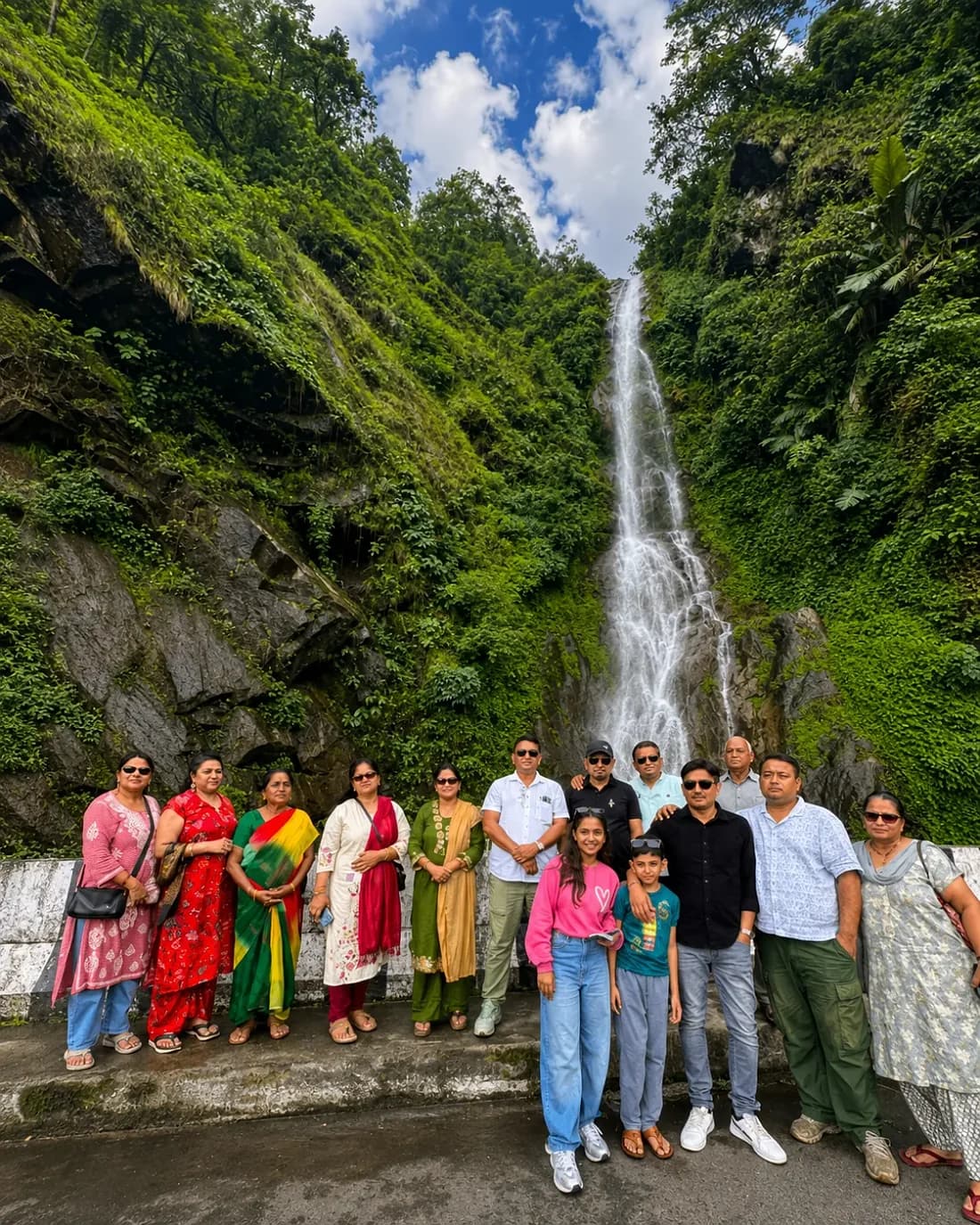 The group at the foot of a tall roadside waterfall in dense green forest