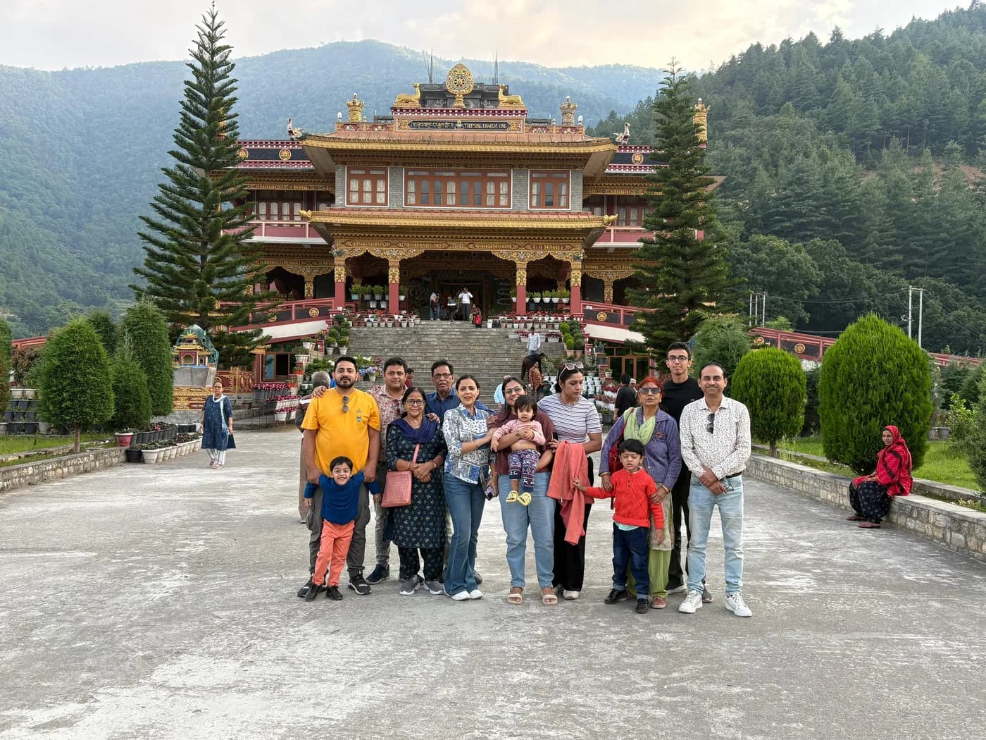 A large family group, children included, on the forecourt of a gold-roofed monastery with wooded hills rising on both sides