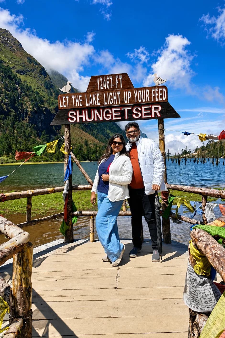 A couple under the Shungetser Lake sign at 12,467 feet, prayer flags and the lake behind them