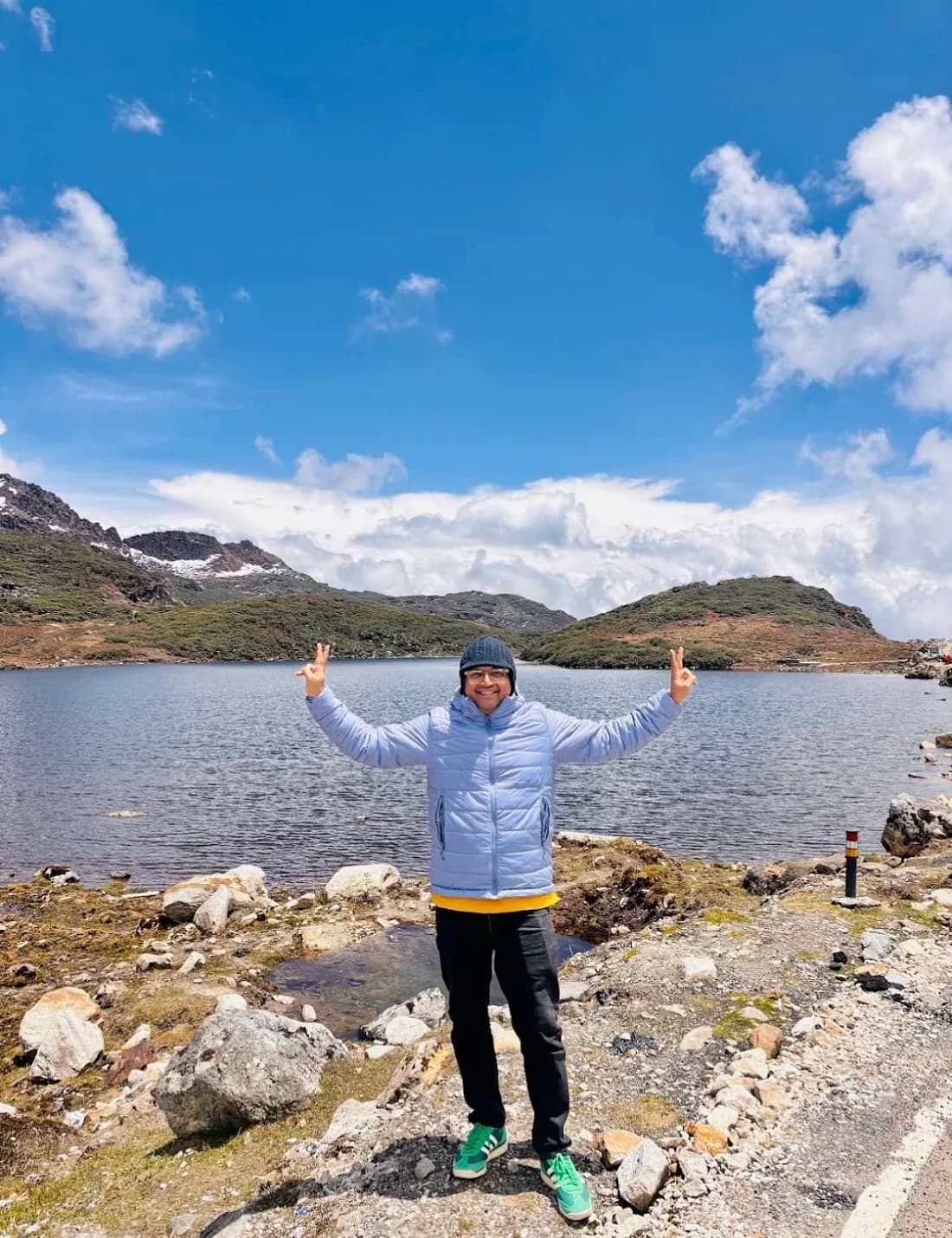 A traveller with both arms raised beside a high lake, snow still lying on the ridges behind