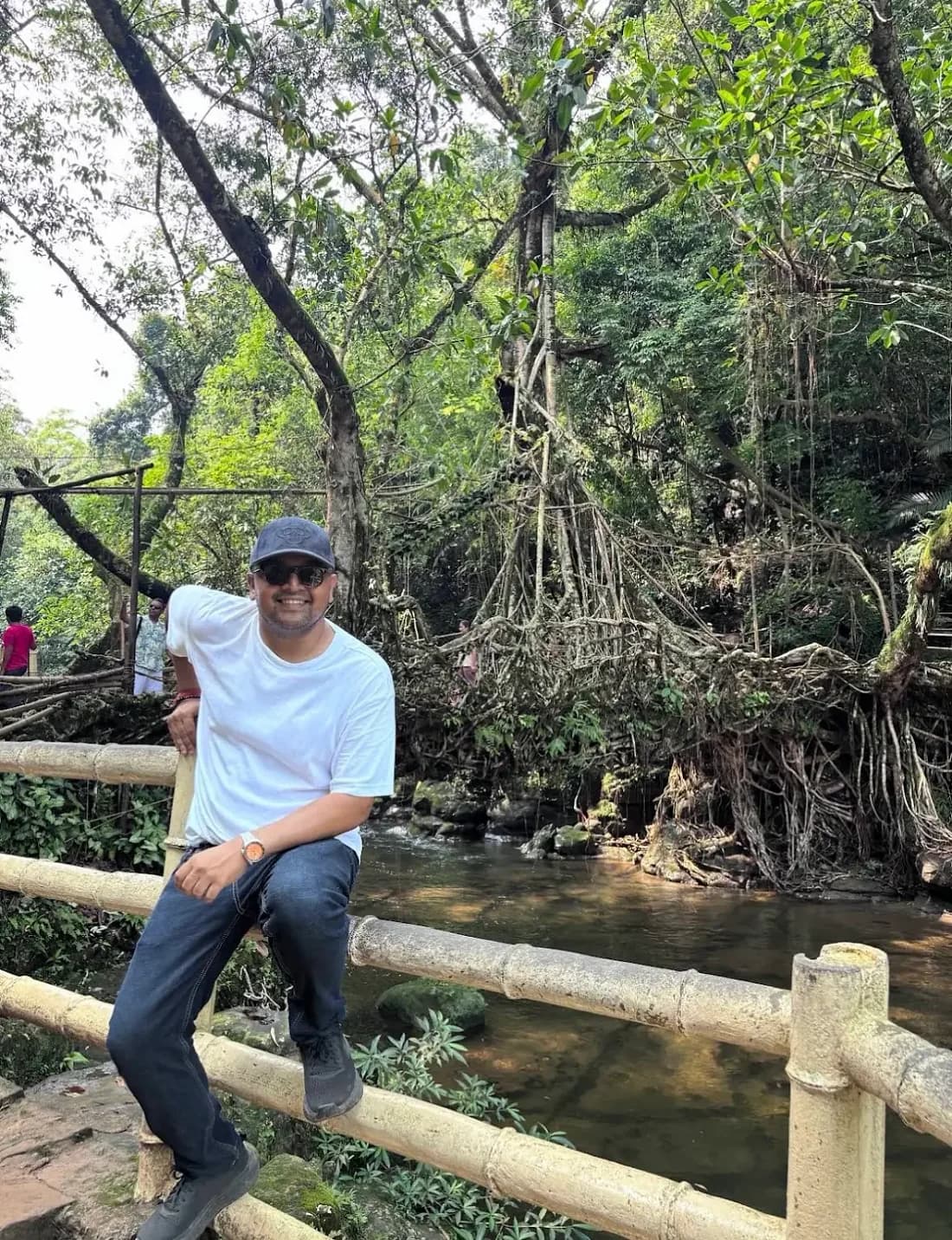 A traveller sitting on a bamboo railing in front of a living root bridge, a stream running below it