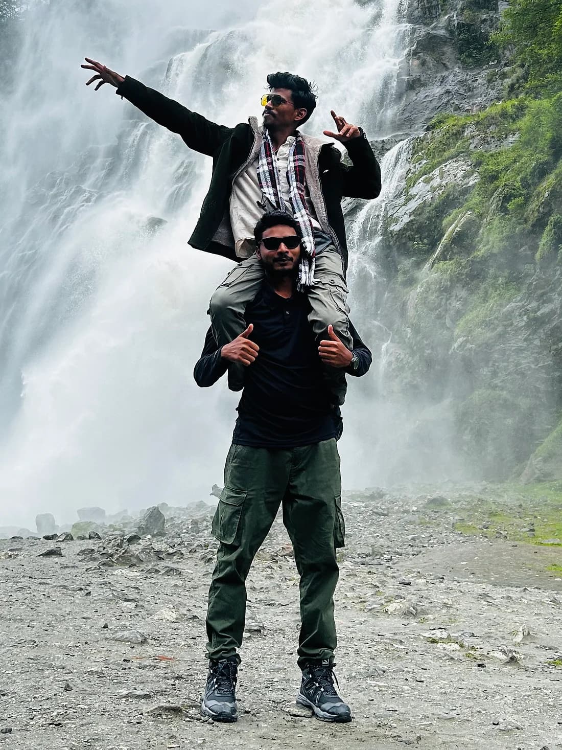 One traveller on another's shoulders in front of the waterfall, arms flung wide