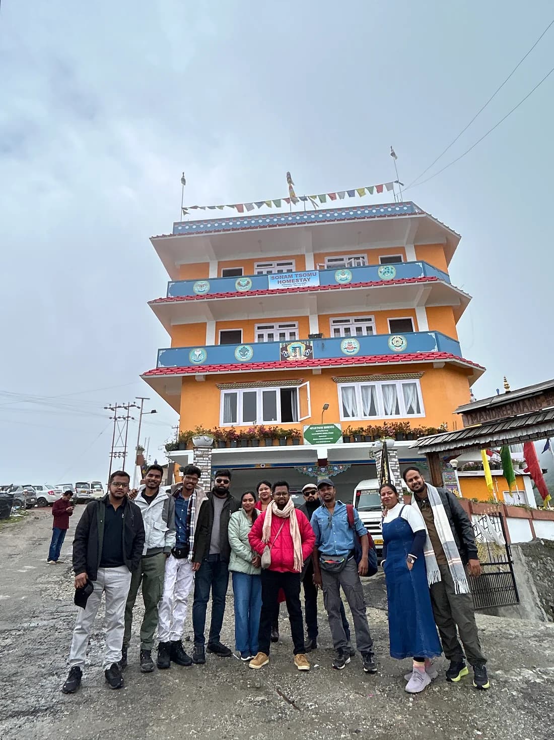 Eleven travellers outside the Sonam Tsomu Homestay, prayer flags along its roofline