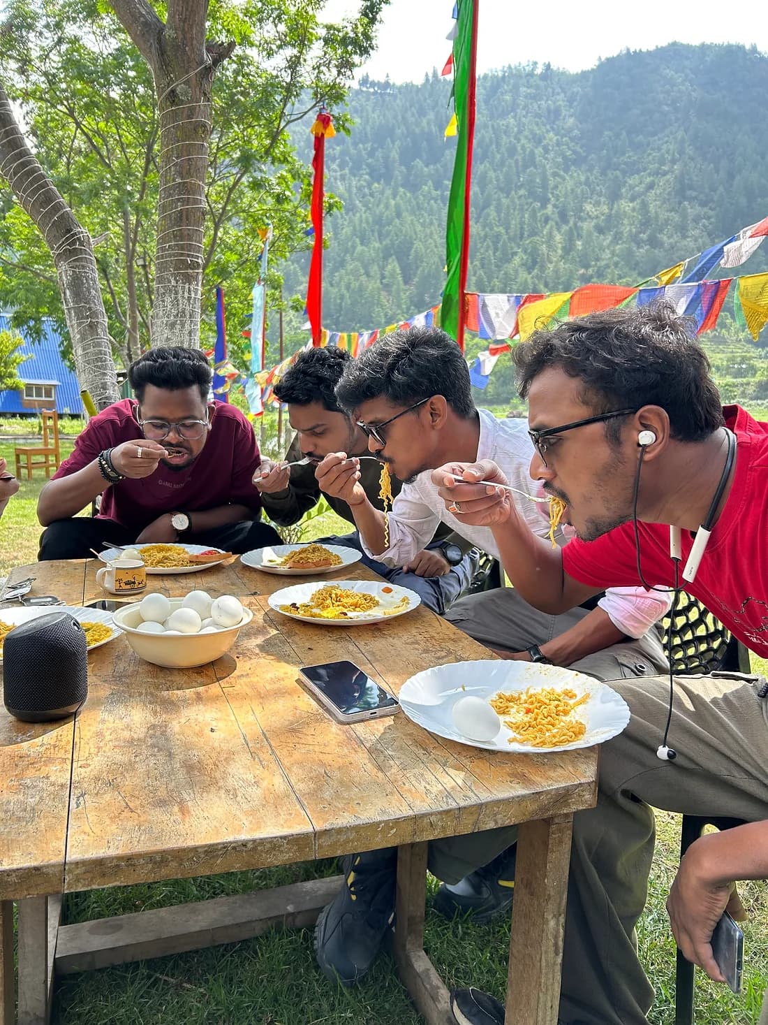 Four travellers eating noodles at a wooden table outdoors, prayer flags in the pines behind