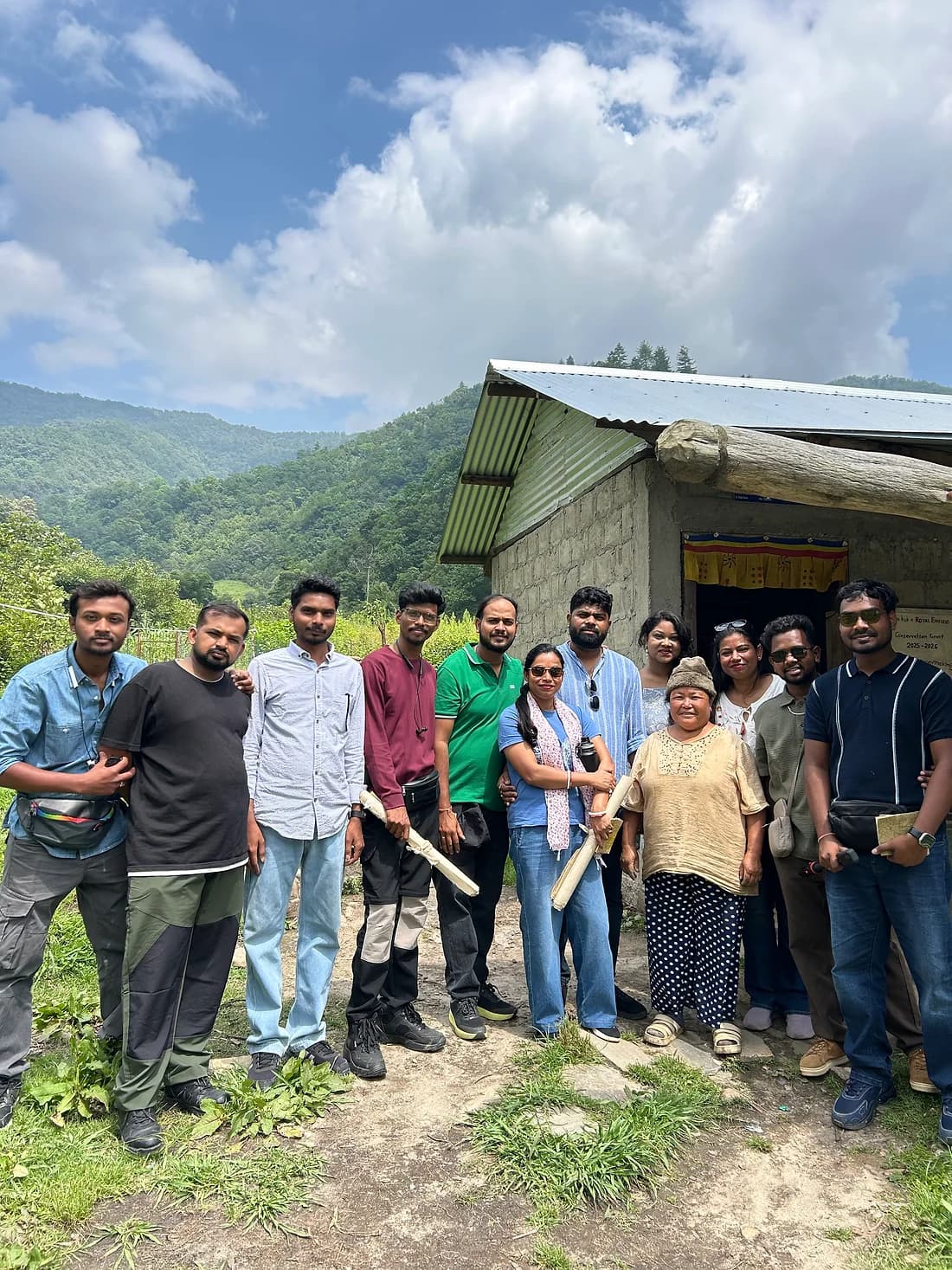 The whole group outside a tin-roofed hut on a green hillside under high cloud