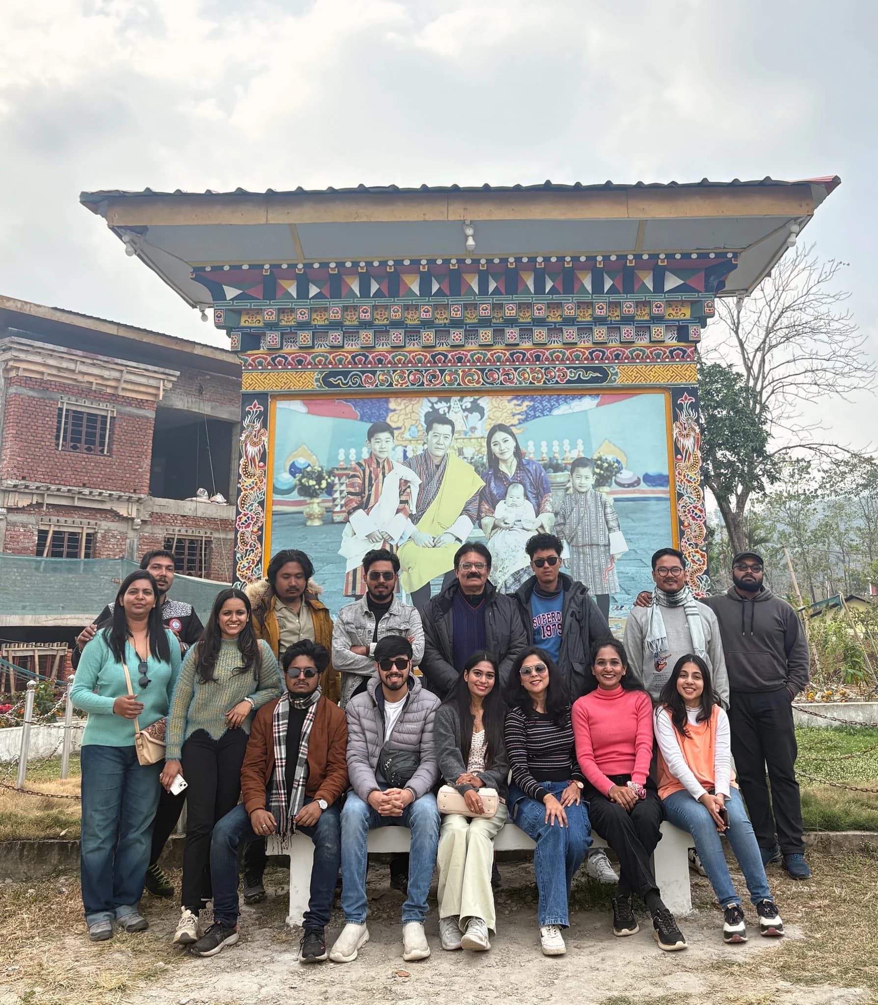 A large group in front of a brightly painted gate under a tiled canopy, a royal portrait filling the panel behind them