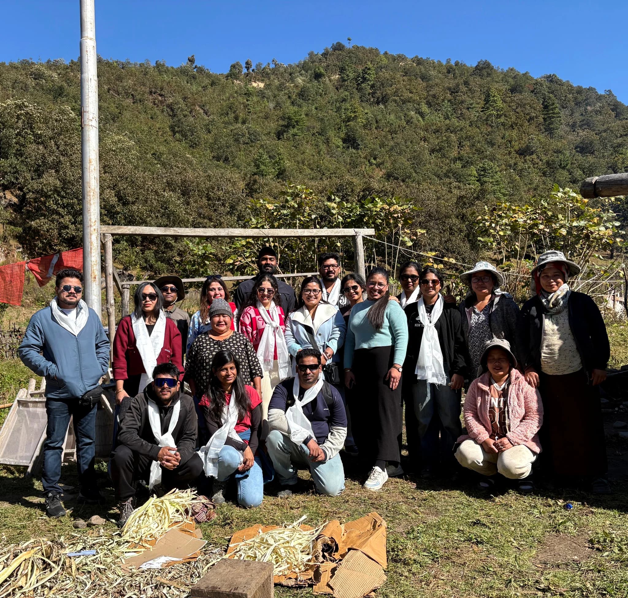 The group and their hosts wearing white ceremonial khada scarves in a hillside clearing, forested slopes behind