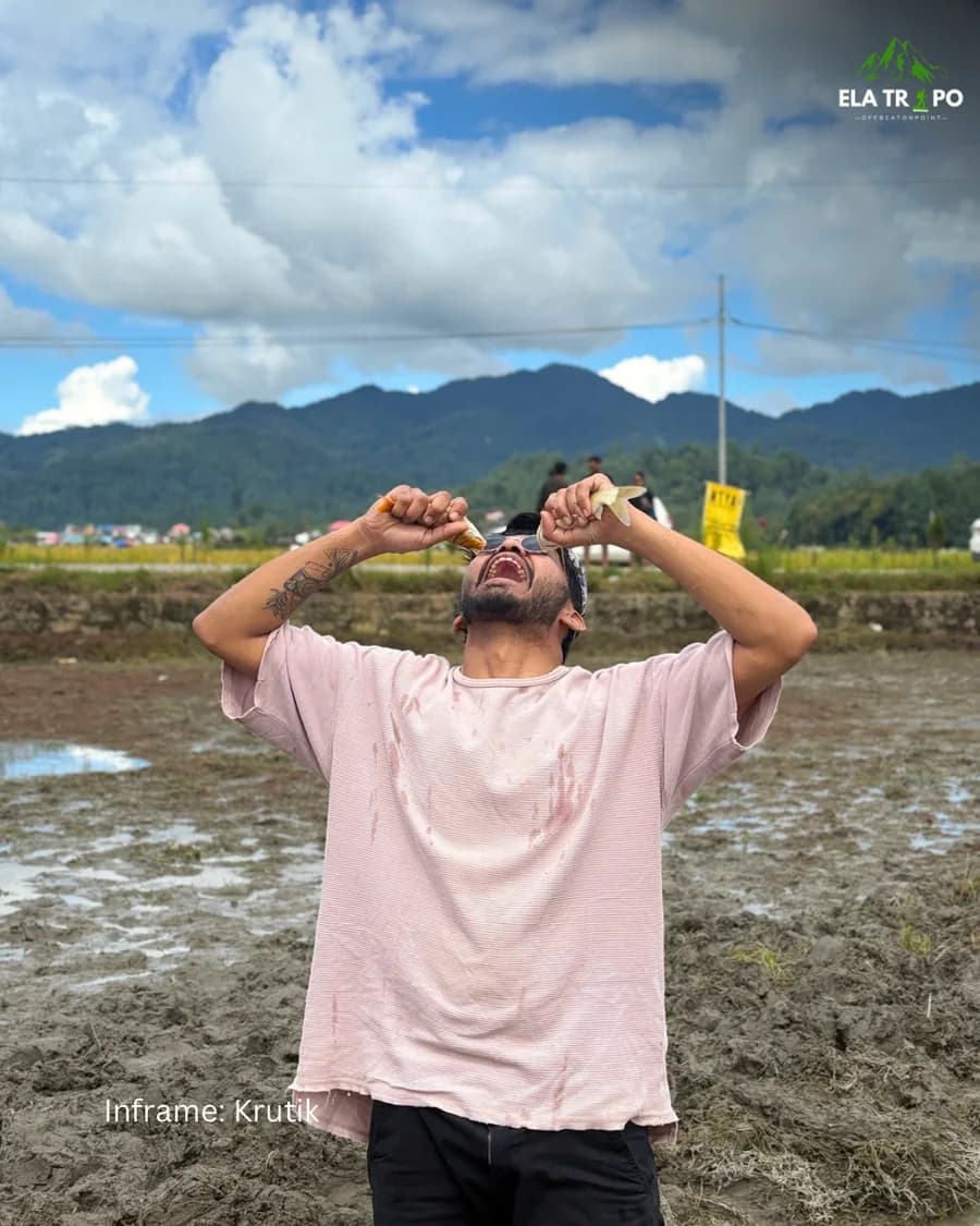 A traveller standing in a flooded paddy field holding up the fish he has just caught, mountains behind