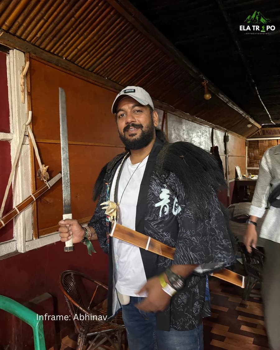 A traveller wearing a traditional feathered coat and holding a dao, traditional stringed instruments on the wall behind him