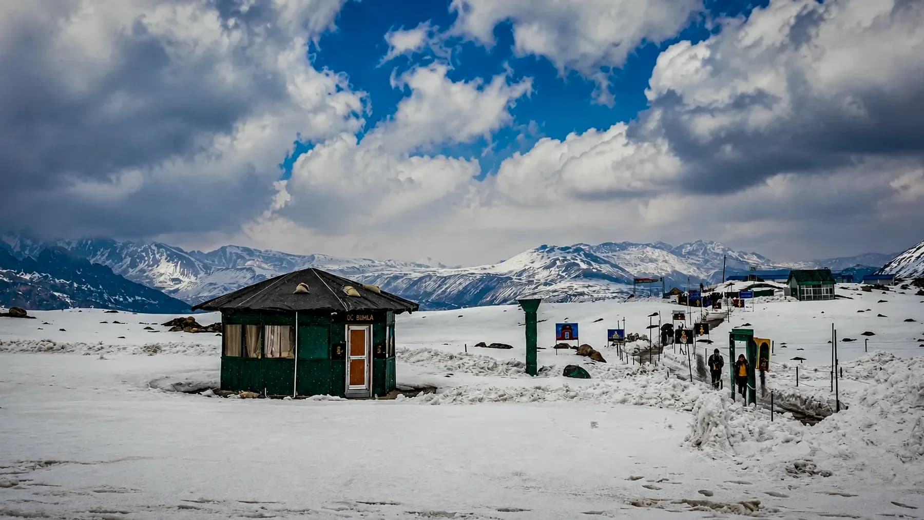 A lone green post hut on a wide snowfield at Bumla, with snow ranges and a bank of cloud behind it