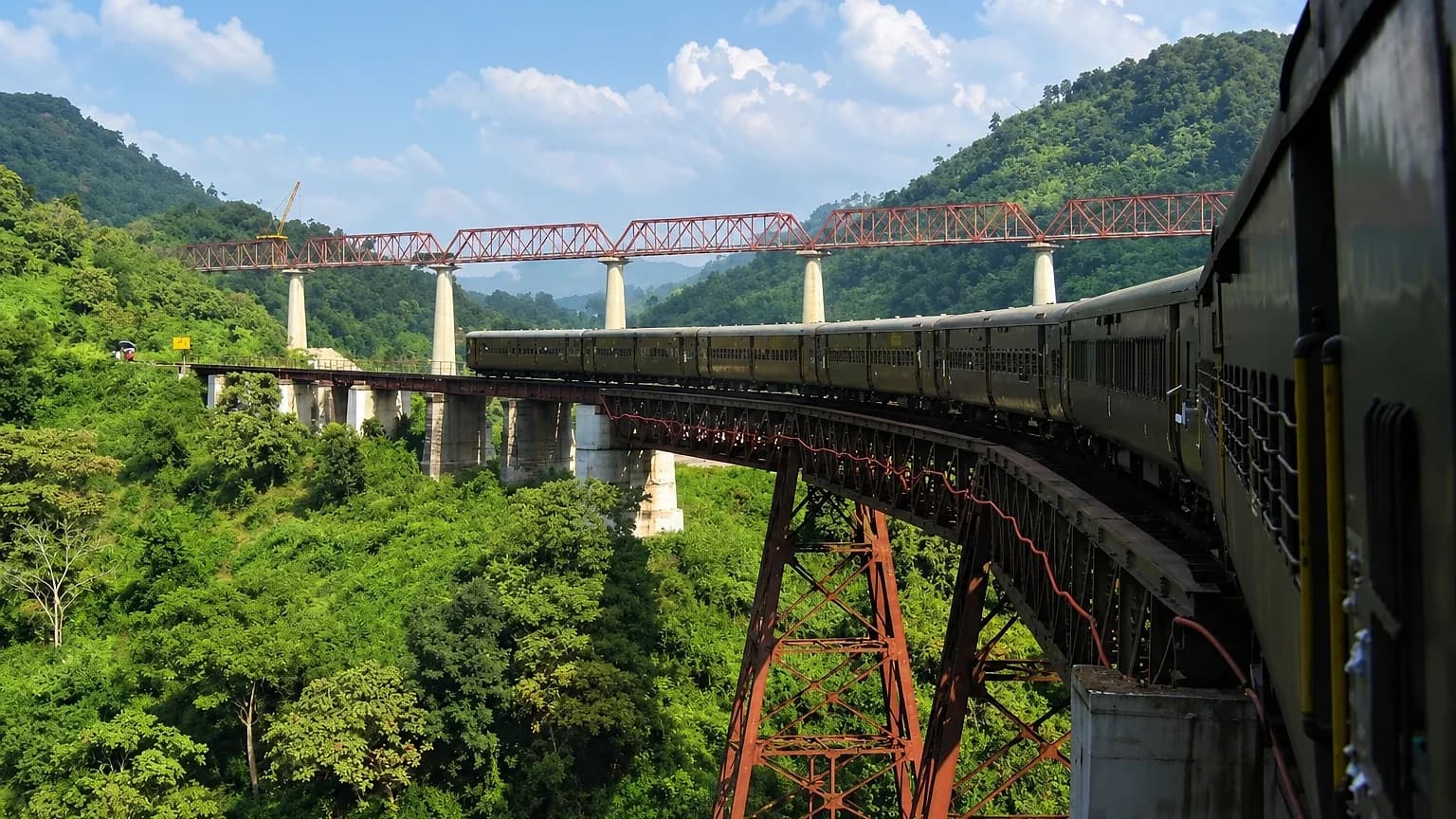 A train crossing a high steel bridge through forested hills on the Haflong line