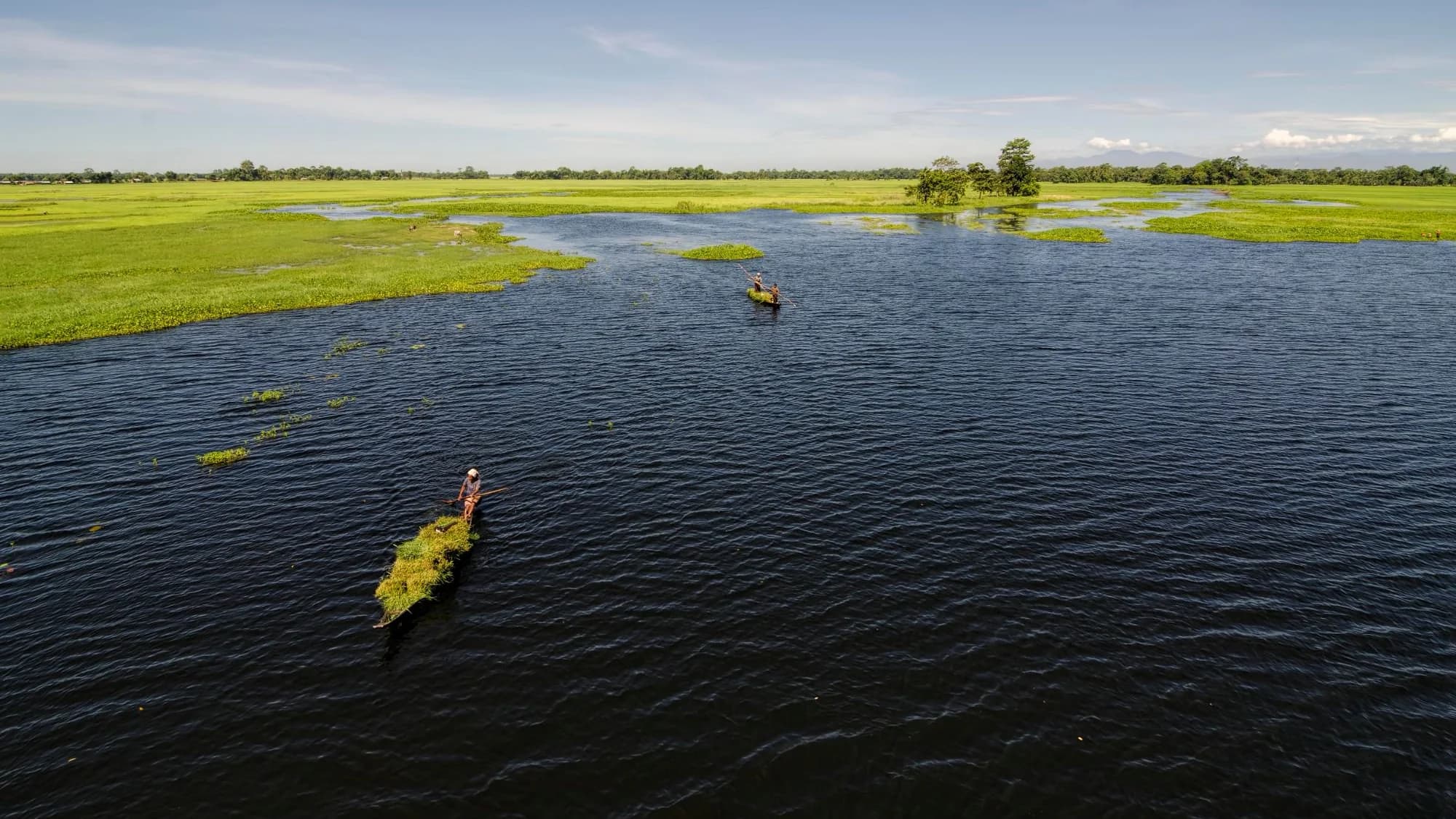 Two poled boats on a wide Brahmaputra channel at Majuli, flat green fields running to the horizon