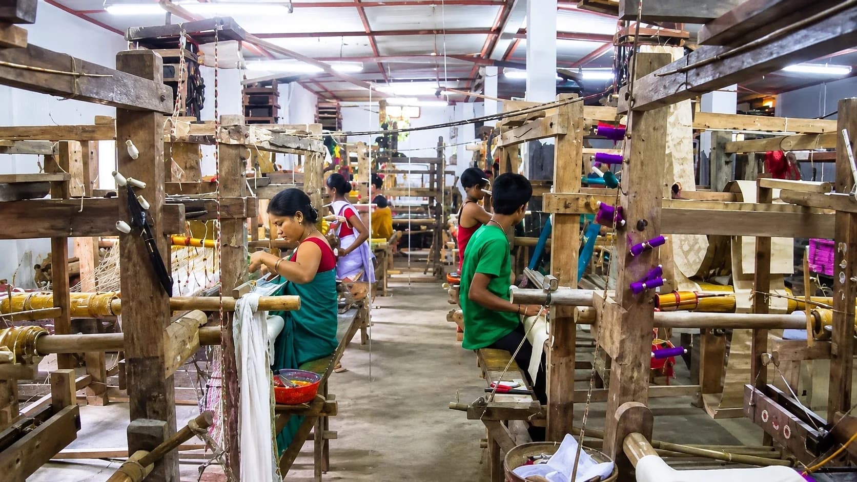 Weavers at wooden handlooms in a long workshop at Sualkuchi, spools of coloured silk above them