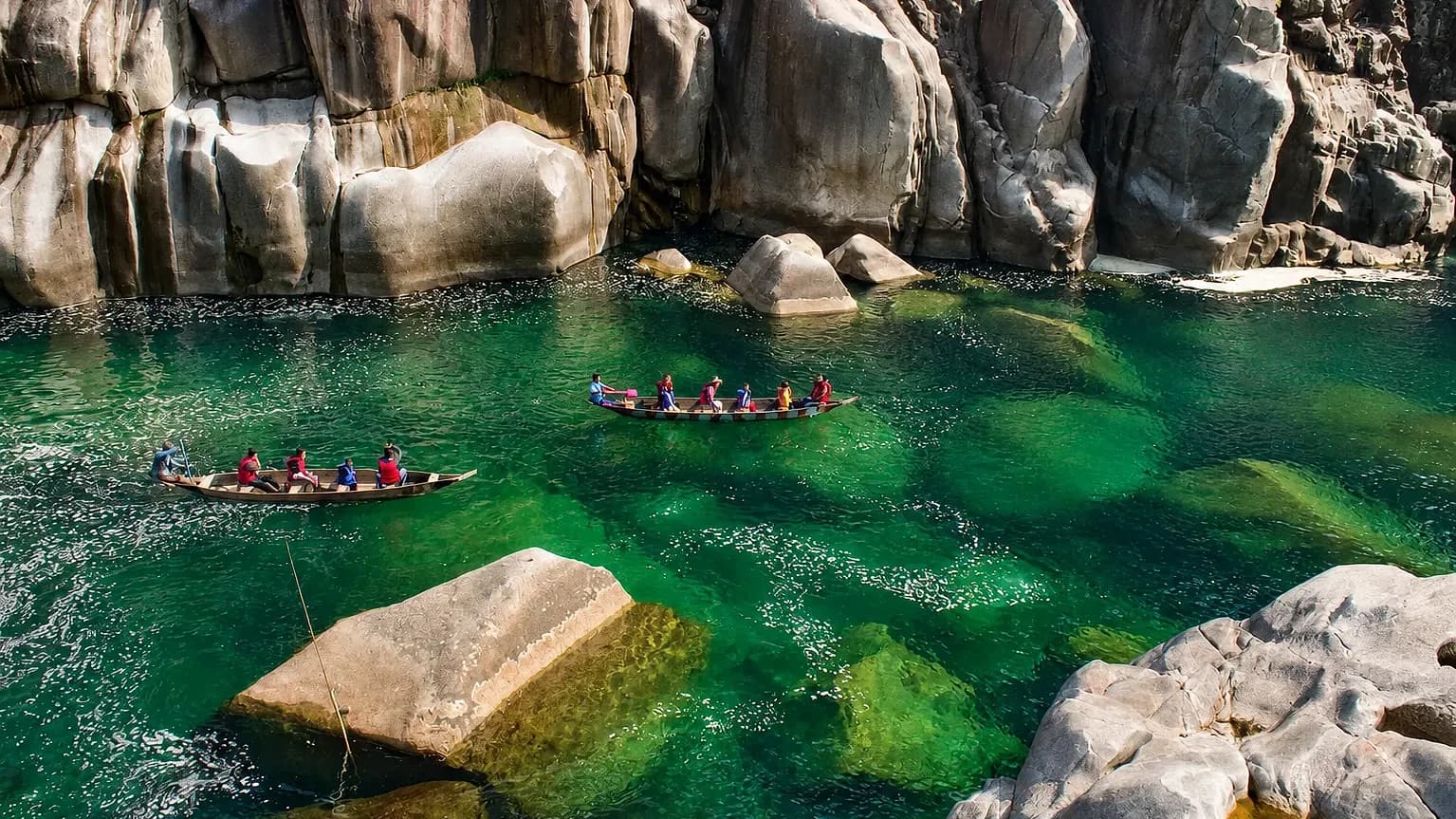 Two boats of paddlers in life jackets on clear green water at Shnongpdeng, pale boulders rising around them
