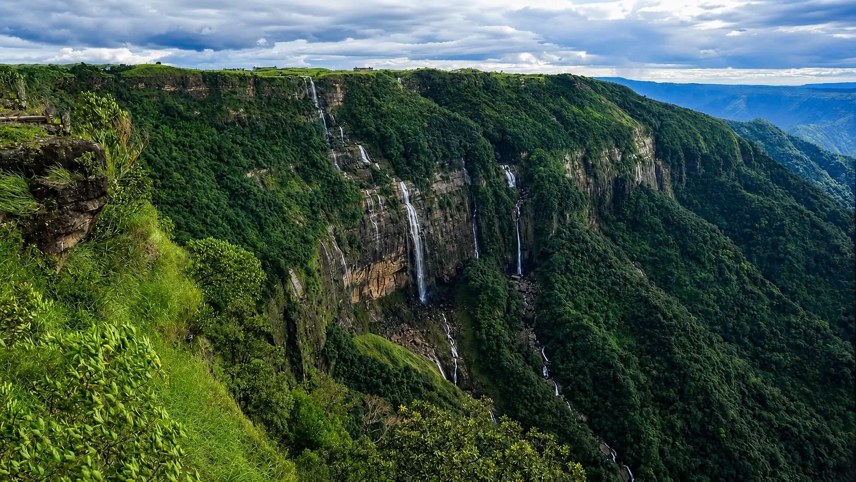 Several waterfalls dropping together off a green plateau edge into a deep forested gorge under heavy cloud