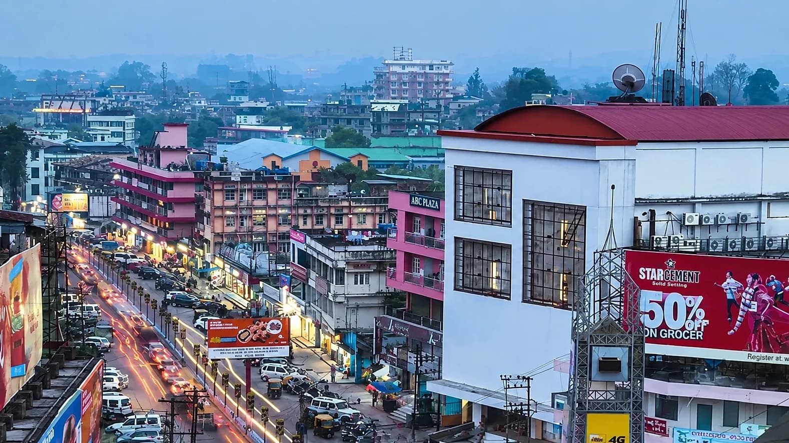 A busy Dimapur street at blue hour, headlights streaking past lit shopfronts and hoardings