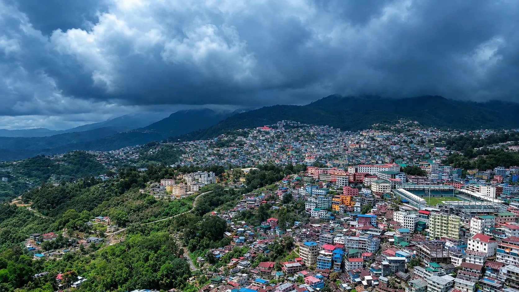 Kohima's coloured rooftops packed across a ridge under heavy monsoon cloud, forested ranges beyond