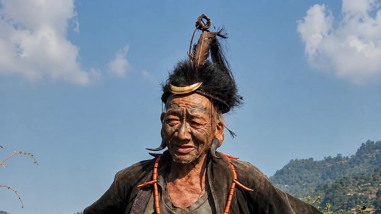 A Konyak elder at Longwa in a feathered headdress and beaded necklace, hills behind him