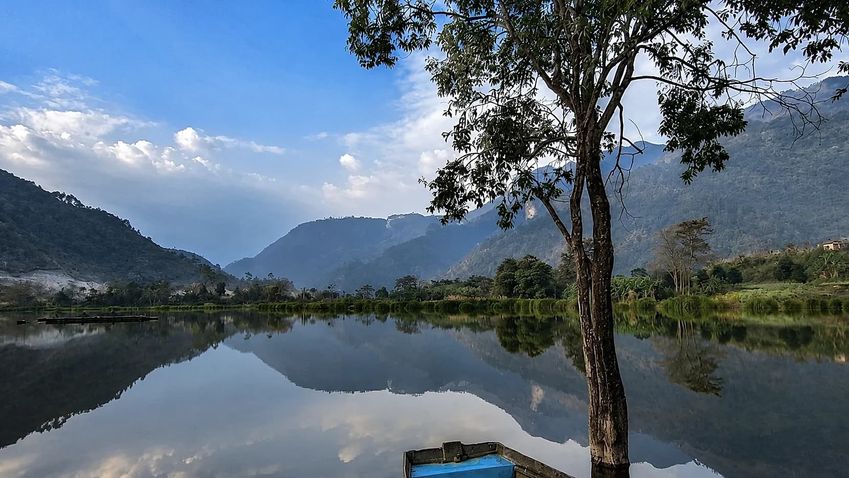 Shilloi Lake lying mirror-still under forested hills, a bare tree and the prow of a boat in the foreground