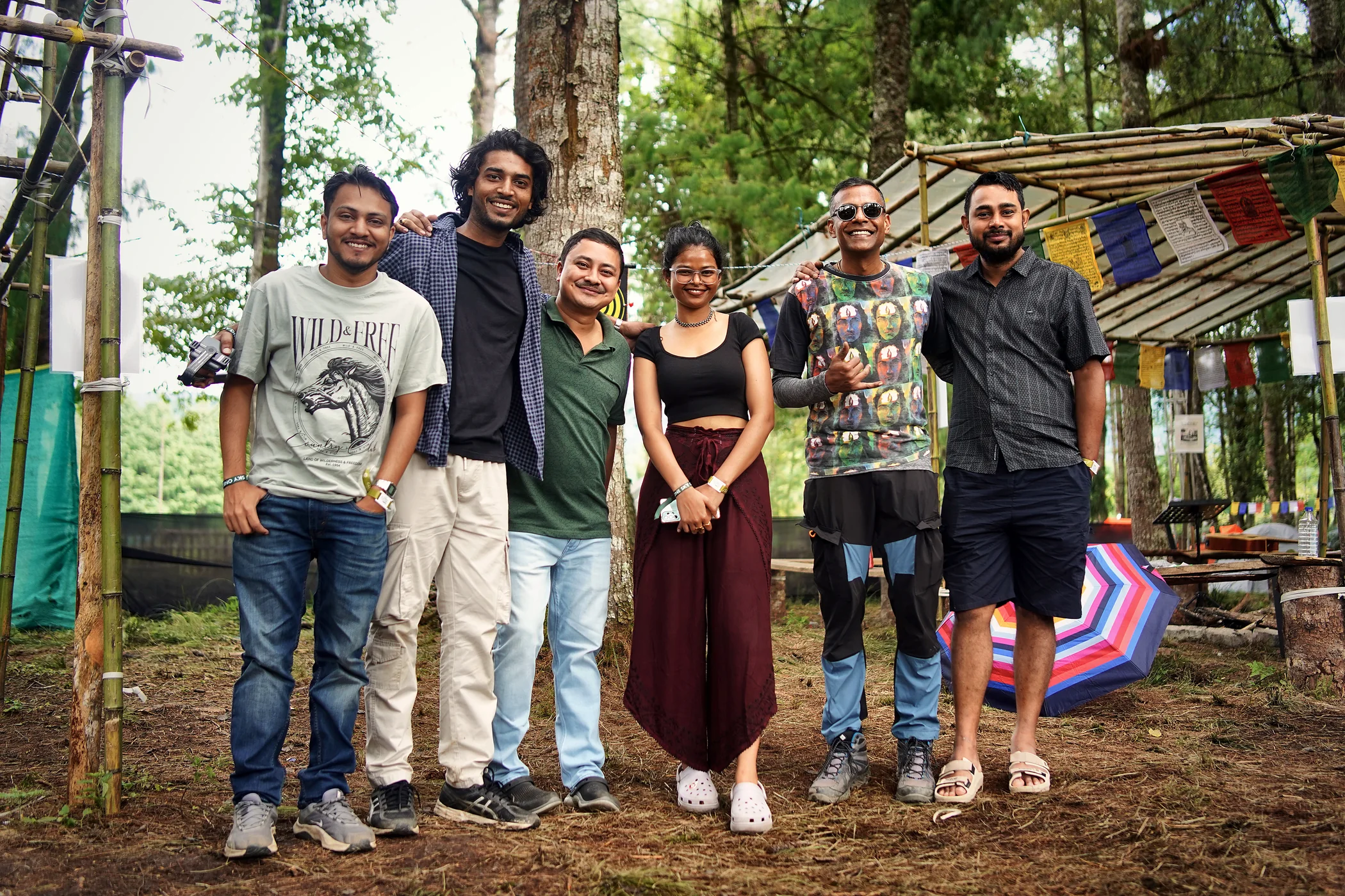 Six campers arm in arm among the pines, prayer flags and a bamboo shelter behind them